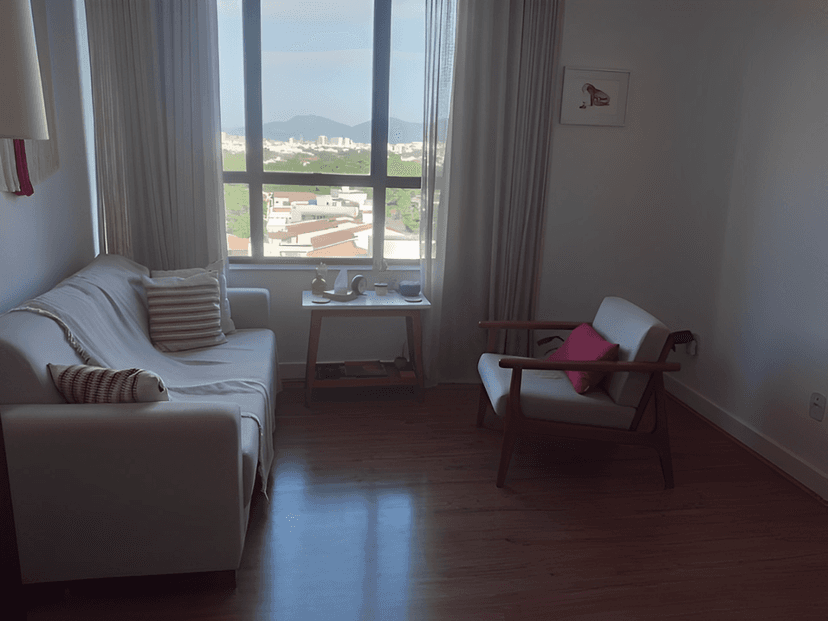 Uma sala de estar com um sofá branco e um assento estofado, um em frente ao outro e ambos na frente de uma janela grande. Há uma mesa de café com uma planta na frente da janela. A sala é iluminada com luz natural que entra pela janela.